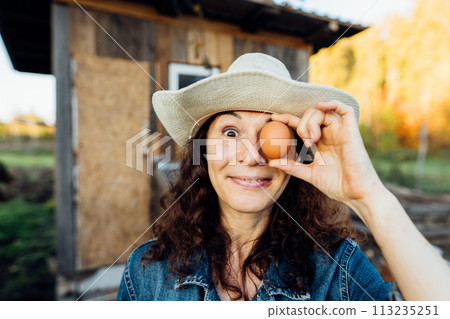 Laughing Woman in Hat Holds Egg to Face, Closing Eye . Fresh chicken egg in the female hands of a farmer, rural life, organic farming Happy moment of collecting eggs Laughing Woman in Hat Holds Egg to Face, Closing Eye . Fresh chicken egg in the female hands of a farmer, rural life, organic farming Happy moment of collecting eggs 113235251