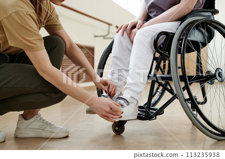 Crop shot of unrecognizable dance coach helping woman in wheelchair to fix shoes on her feet 113235938