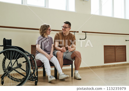 Long shot of cheerful young female wheelchair dancer sitting on bench chatting with her male trainer during break 113235979