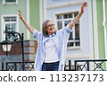 A woman standing in front of a building, arms outstretched. 113237173