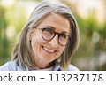 An older woman wearing glasses smiles directly at the camera. 113237178
