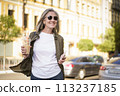 A woman strolling along a busy street while carrying a beverage in her hand. 113237185