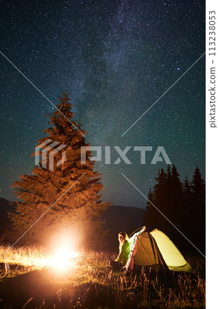 Night camping in mountains under starry sky and Milky way. Female tourist sitting in tent entrance in campsite, admiring landscape and burning campfire. Concept of traveling and hiking. Night camping in mountains under starry sky and Milky way. Female tourist sitting in tent entrance in campsite, admiring landscape and burning campfire. Concept of traveling and hiking. 113238053