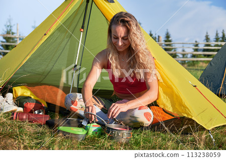 Woman tourist camping in mountains. Sporty woman traveling, hiking in summer. Attractive, young female sitting on grass near tent, cooking. Concept of harmony with nature. 113238059