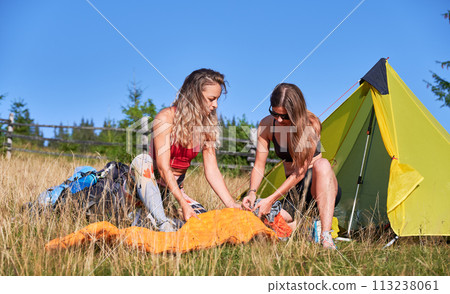Two women travellers camping outdoors. Female tourists inflating the air mattress, wearing sportswear, hiking in mountains, making campsite. Concept of tourism and traveling. 113238061