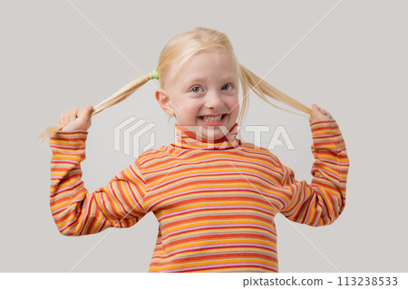 Portrait of a cute little girl with blond hair holding her pigtails on a gray background Portrait of a cute little girl with blond hair holding her pigtails on a gray background 113238533