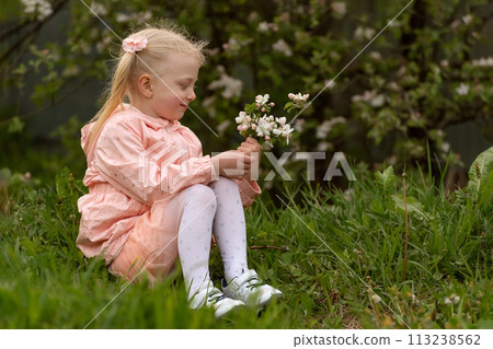 Smiling little lady in pink dress in the garden. Girl sitting with blooming branch and looking on their 113238562