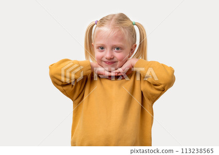 Portrait of a little girl with blond hair in a yellow sweatshirt holds her hands to her chin. Portrait of a little girl with blond hair in a yellow sweatshirt holds her hands to her chin. 113238565