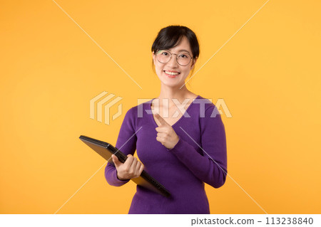 A confident young woman, wearing a purple shirt and eyeglasses, holds a laptop against a vibrant yellow background, symbolizing success and professionalism in a studio portrait. 113238840