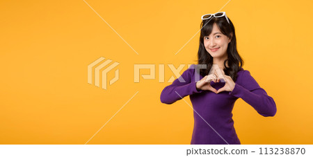 Young cheerful asian woman 30s wearing purple sweater dress and sunglasses shapes heart gesture on yellow background. 113238870