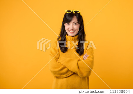An attractive young woman in her 30s wearing yellow sweater and sunglasses, confidently posing with crossed arms isolated on yellow background, representing success and a positive business lifestyle. 113238904