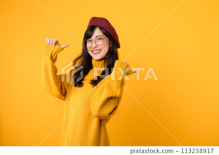 Join the promo fun with a stylish woman, wearing a red beret and yellow sweater, as she points her thumb to free copy space against a vibrant yellow background. 113238917