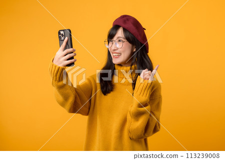 A stylish woman in a red beret and yellow sweater poses with her smartphone, capturing the moment against a vibrant yellow background. A stylish woman in a red beret and yellow sweater poses with her smartphone, capturing the moment against a vibrant yellow background. 113239008