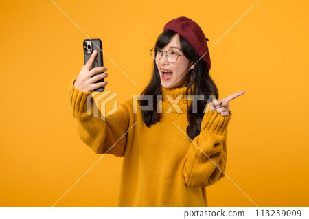 Portrait young beautiful 30s asian woman happy smile wearing yellow shirt and red hat pointing finger gesture to free copy space and using smartphone against yellow background. new application 113239009