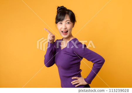 Portrait cheerful young asian woman 30s wearing purple shirt pointing finger to free copy space isolated on yellow background. Portrait cheerful young asian woman 30s wearing purple shirt pointing finger to free copy space isolated on yellow background. 113239028