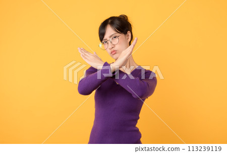 Explore concept of denial and rejection through X gesture made serious young woman wearing purple shirt and eyeglasses in studio portrait against a yellow background. An impactful symbol for caution. 113239119