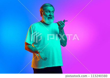 Emotional bearded senior man recording voice message via mobile phone on gradient blue-purple background in neon light 113240380