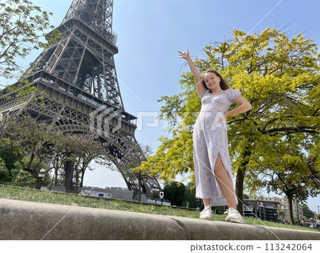 a cheerful teen girl stands against the backdrop of the Eiffel Tower in a beautiful dress, she raised her hand, you can advertise a travel agency trip to Paris France 113242064