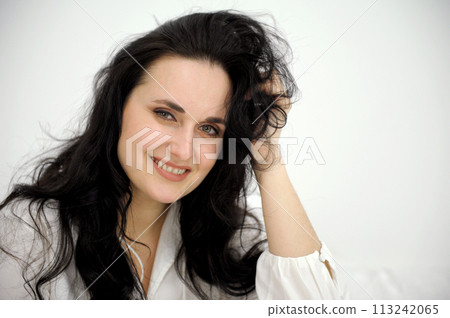 beautiful brunette woman straightens her hair and gently smiles looks bright eyes white background studio on white sheets bed rest relax beautiful face red manicure make eyes flirt 113242065