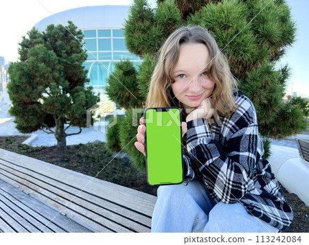 A young woman sits with a phone turned to the camera with a green screen Vancouver Porto Canada Place Coniferous trees are in the background. She is wearing a black and white checkered blouse. 113242084