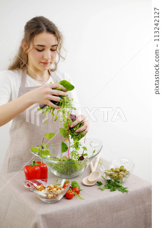 young girl mixes lettuce with hands she pours into glass plate vegetarian food wooden spoons apron tablecloth white background young woman teenager prepares dinner for family dinner breakfast lunch 113242127