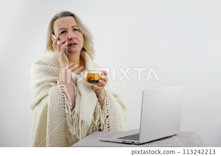 View over the shoulders of a sick couple in hats and blankets using a laptop for an online conference with their doctor. woman with a fever are warmed by tea and have their temperature. View over the shoulders of a sick couple in hats and blankets using a laptop for an online conference with their doctor. woman with a fever are warmed by tea and have their temperature. 113242213