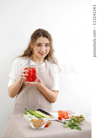 Smiling brunette woman holding red bell pepper while standing with hand on hip Green vegan salad with mixed green leaves and vegetables Green salad with tomatoes and fresh vegetables 113242261