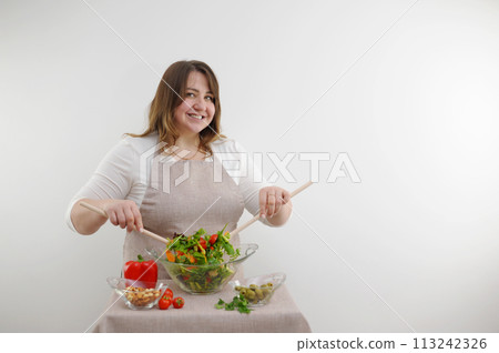 successful promotional photo of beautiful curvy woman preparing salad diet for weight loss beige apron Same color tablecloth wooden spoons vegetables slicing ingredients for delicious fresh salad 113242326