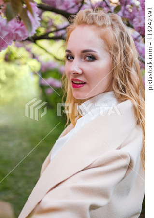 Red-haired girl with pink lipstick stands near pink flowering tree looks into the frame and smiles. Attractive young woman. Vertical frame. Red-haired girl with pink lipstick stands near pink flowering tree looks into the frame and smiles. Attractive young woman. Vertical frame. 113242591
