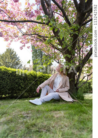 Young woman sitting on lawn in blossoming park, warm spring day. Attractive girl on rest. Vertical frame. Young woman sitting on lawn in blossoming park, warm spring day. Attractive girl on rest. Vertical frame. 113242592