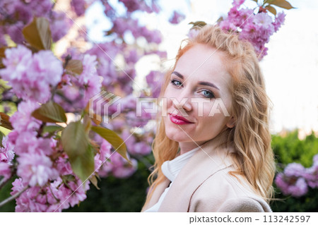Portrait of happy attractive woman in beige coat among blossoming sakura branches. Stylish girl with red lips in blooming park. Portrait of happy attractive woman in beige coat among blossoming sakura branches. Stylish girl with red lips in blooming park. 113242597