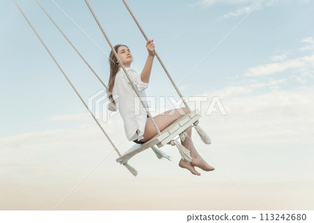 Young beautiful girl in white shirt on rope swing high on blue sky background during a sunset time. 113242680