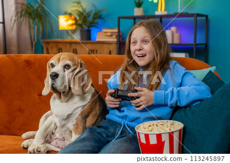 Girl child using joystick controller playing video console television game sits on sofa in room 113245897