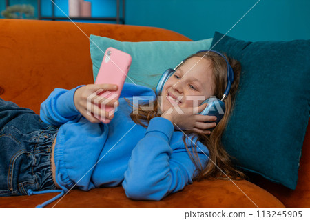 Happy Caucasian girl child in wireless headphones relaxing lying on couch at home listening music Happy Caucasian girl child in wireless headphones relaxing lying on couch at home listening music 113245905