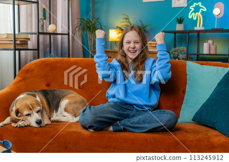 Cheerful young child girl celebrating victory winning the lottery at home beside beagle dog on sofa 113245912