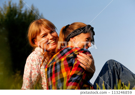 A heartwarming portrait of a mother and daughter sharing a tender cuddle outdoors on a sunny summer day, radiating happiness and familial love, perfect for illustrating family bonding moments A heartwarming portrait of a mother and daughter sharing a tender cuddle outdoors on a sunny summer day, radiating happiness and familial love, perfect for illustrating family bonding moments 113246859