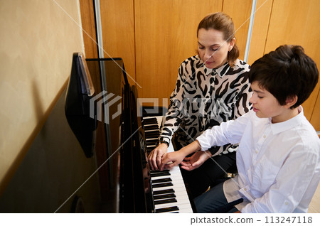 Authentic portrait of a woman pianist musician holding the hands of a teenage boy, showing the true position of finger on piano keys, explaining the piano lesson during individual music class at home 113247118