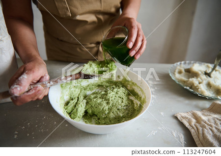 Close-up hands of a housewife, chef pastry pouring green spinach water into a bowl with wheat four, making dough of green color for ravioli or dumplings with mashed potato, in the rustic home kitchen Close-up hands of a housewife, chef pastry pouring green spinach water into a bowl with wheat four, making dough of green color for ravioli or dumplings with mashed potato, in the rustic home kitchen 113247604