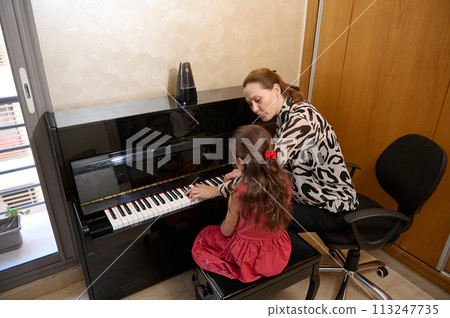Rear view of a cute little girl in elegant red dress, sitting at wooden piano, with teacher learning to play piano indoors. Musical education and talent development in progress 113247735