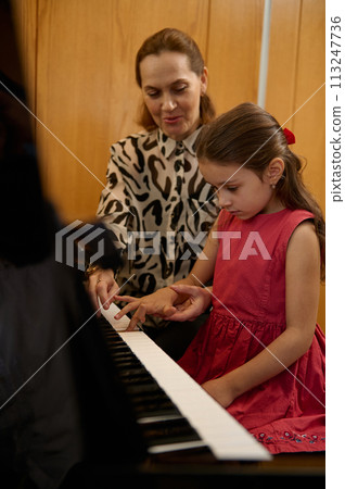 Vertical closeup portrait of a cute little girl in elegant red dress, sitting at wooden piano, with teacher learning to play piano indoors Vertical closeup portrait of a cute little girl in elegant red dress, sitting at wooden piano, with teacher learning to play piano indoors 113247736