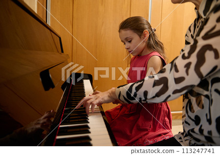 Little girl having a piano lesson with her teacher at home. Little child girl putting her fingers on black and white keys, creating the rhythm of music, composing melody and singing Christmas song 113247741