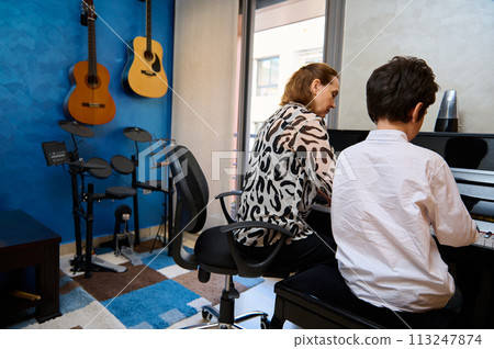 Rear view of a teenage student boy playing piano, composing a melody under his teacher's guidance during individual music lesson at home. Children. Education. Lifestyles. Hobbies and entertainment. 113247874