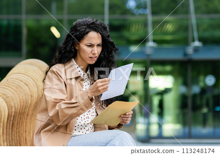 A worried young woman is sitting on a bench outside, intently reading a document with a serious expression. A worried young woman is sitting on a bench outside, intently reading a document with a serious expression. 113248174