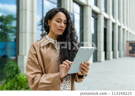 Confident professional woman browsing on a digital tablet while standing outside a contemporary office building. 113248225