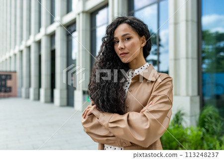 A young professional woman stands confidently outside a modern office building, exuding strength and ambition in a corporate setting. 113248237