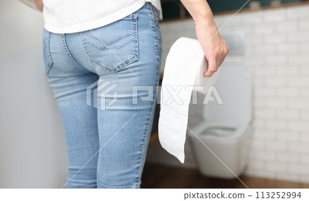 Woman in blue jeans stands with back and holds roll of toilet paper in her hand in front of toilet close-up 113252994