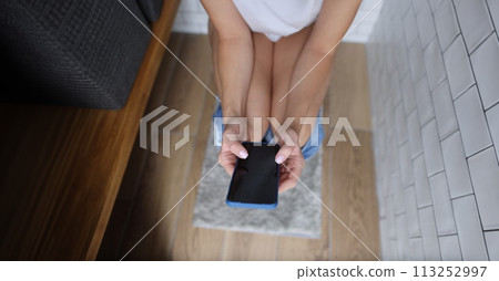 Woman holds phone in her hands having pulled down jeans on toilet in bathroom close-up 113252997
