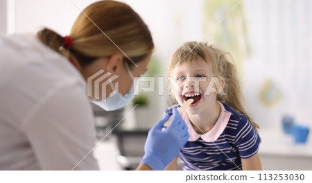 Woman doctor pediatrician in protective medical mask and rubber gloves examines throat of little girl wooden spatula portrait 113253030