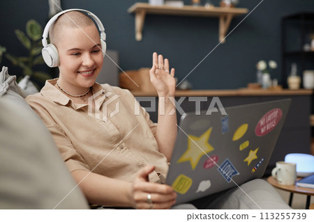 Cheerful young Caucasian woman with shaved head wearing headphones greeting her friend on video call on laptop 113255739