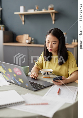 Vertical shot of young Asian female university student sitting at table in morning having breakfast and using laptop 113255753